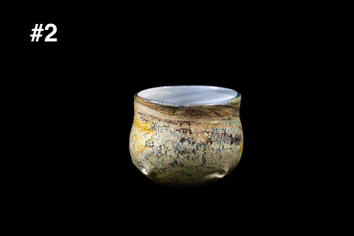 Rounded handmade glass cup with mottled gold, blue, and smoky gray coloring and a gently flared rim against a black background