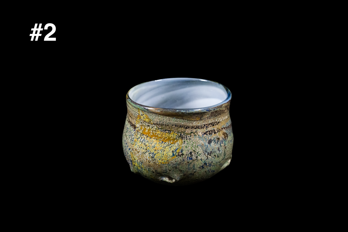 Handmade glass cup with a rounded shape, mottled gold, blue, and gray surface, and a milky white interior against a black background