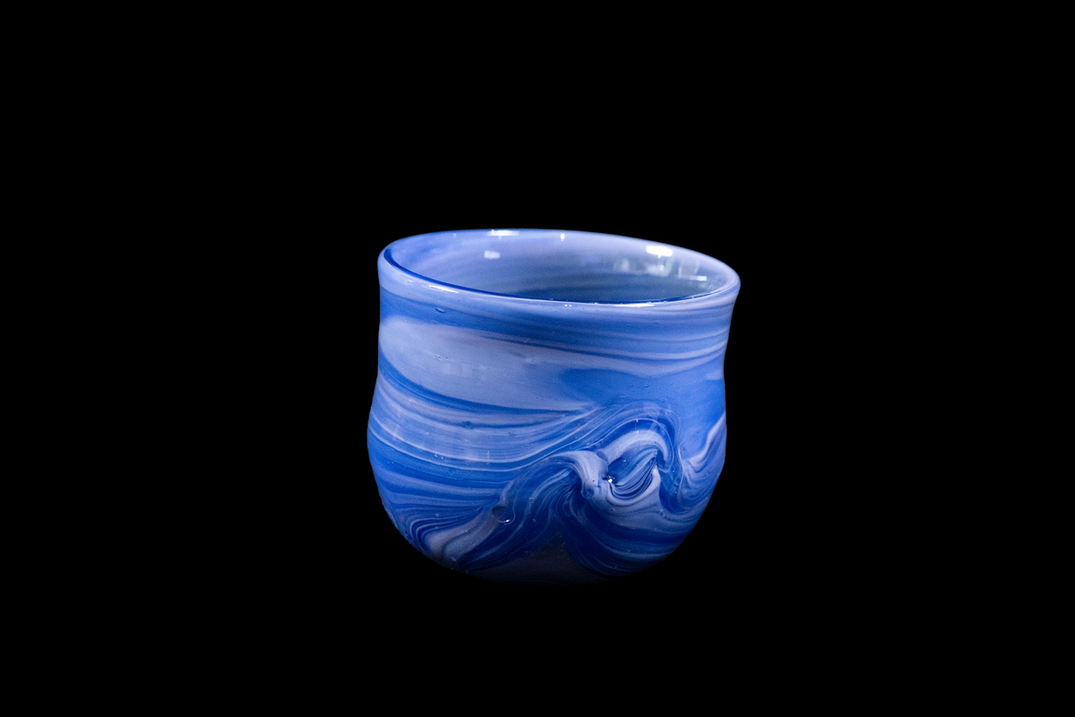 Blue and white marbled glass cup with layered flowing swirls, a rounded rim, and sculpted wave patterns along the lower sides against a black background