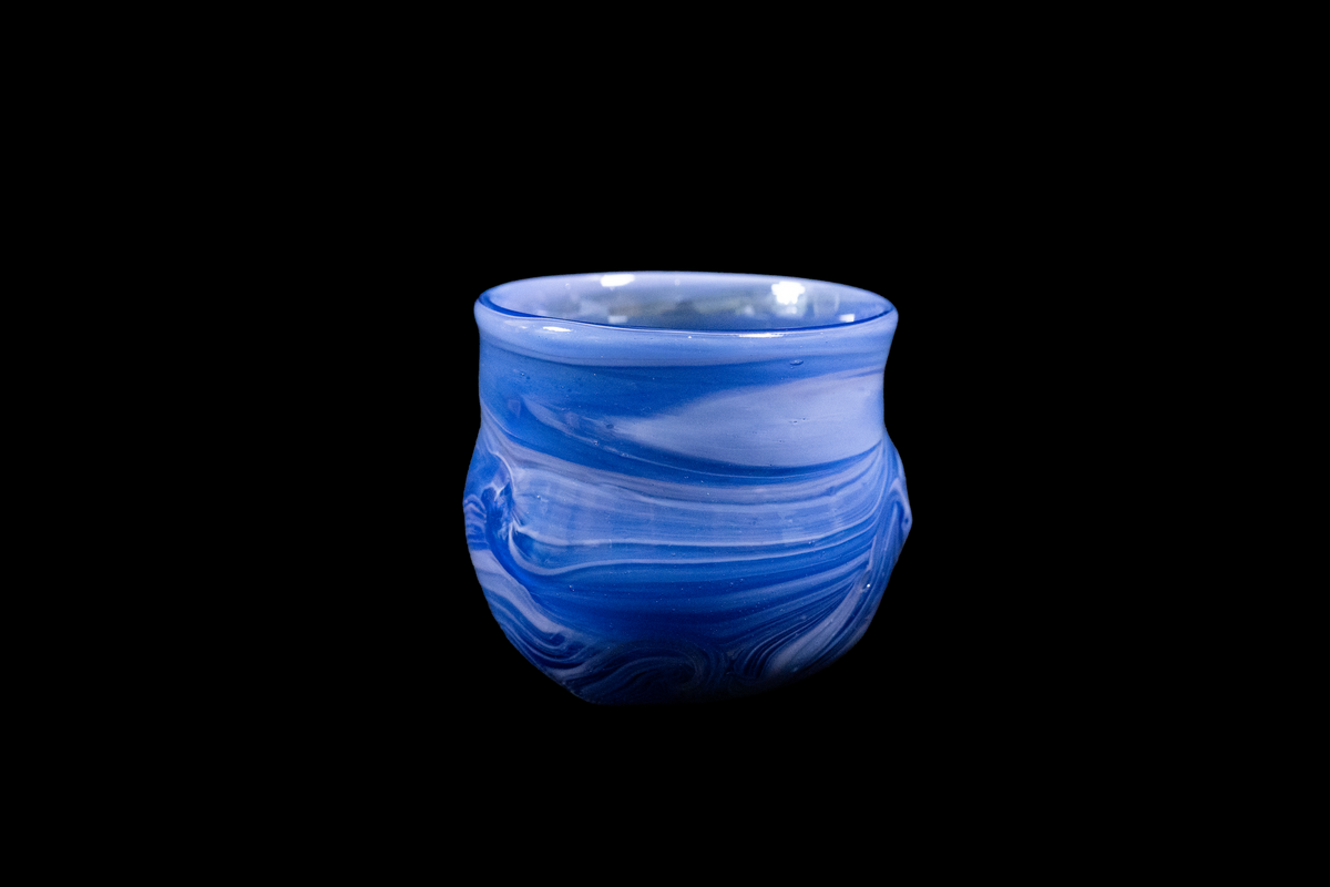 Handmade blue glass cup with layered flowing swirls, rounded sides, and a gently flared rim against a black background