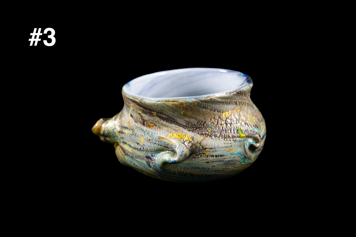 Handmade glass cup with a squat rounded form, milky white interior, and shimmering gold, blue, and green swirls beneath an uneven rim against a black background