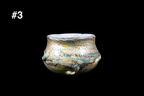 Handmade glass drinking cup with a rounded sculptural form and flared rim featuring shimmering silver blue gold and green swirls on a dark background