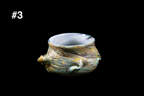 Handmade glass cup with a wide opening, a sculpted shell-like knob on one side, and swirling gold, blue, and silver textures against a black background