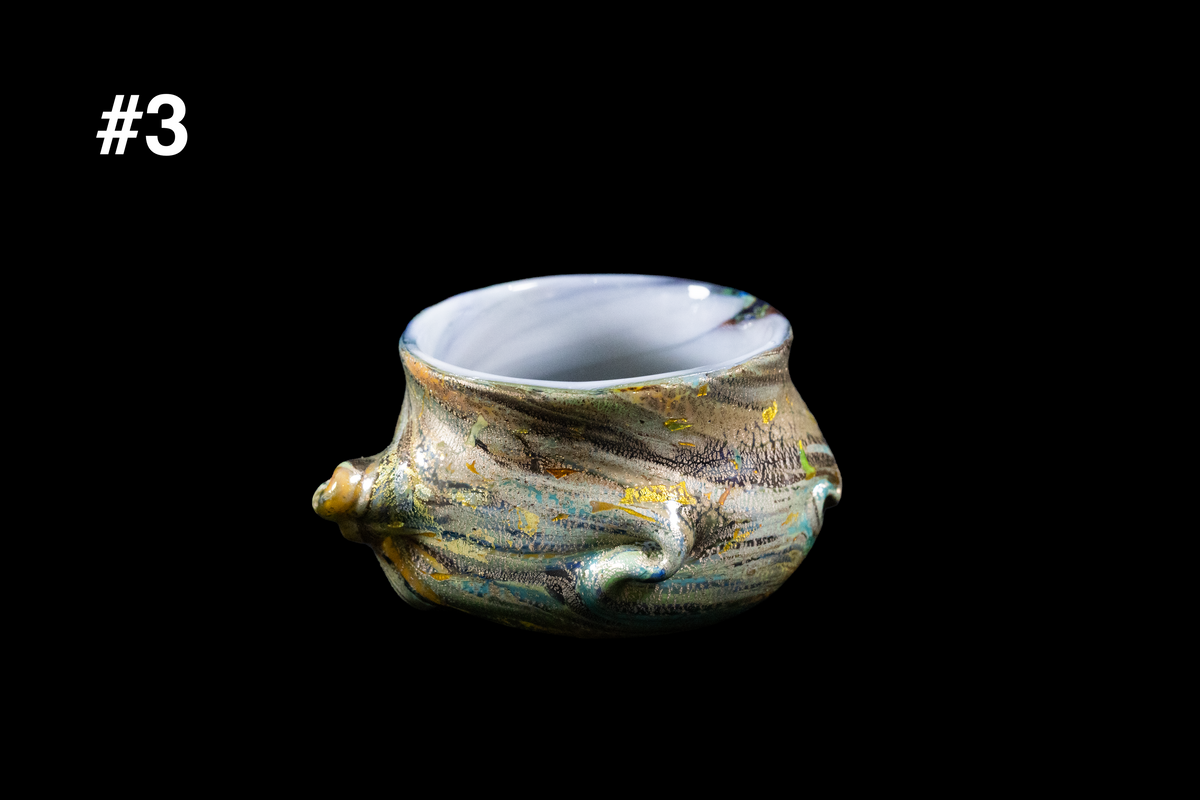 Handmade glass cup with a wide opening, a sculpted shell-like knob on one side, and swirling gold, blue, and silver textures against a black background