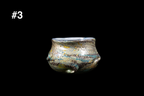 Handmade glass cup with a squat rounded form, sculpted wave accents, and shimmering blue, gold, gray, and black swirls against a black background
