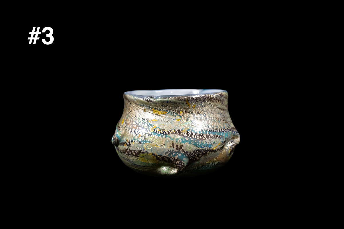 Handmade glass cup with a squat rounded form, sculpted wave accents, and shimmering blue, gold, gray, and black swirls against a black background