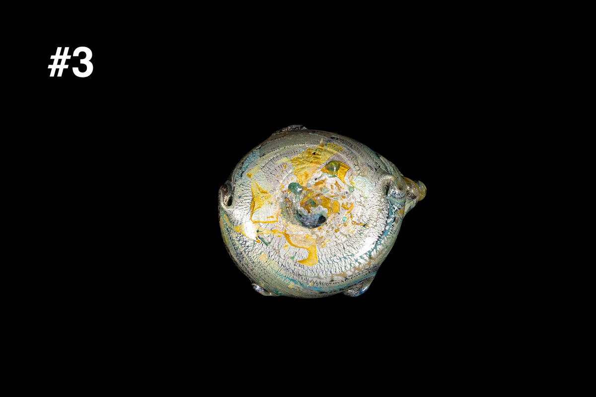 Bottom view of a handmade glass cup with shimmering gold, blue, and silver swirls and sculpted knobs around the base against a black background