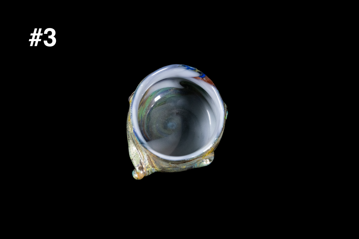 Small handmade glass drinking cup with blue green and smoky gray swirls inside an organically shaped rim against a black background