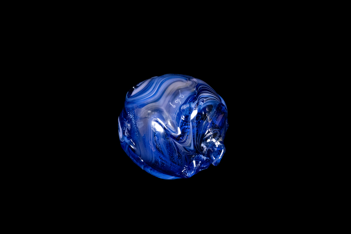 Handmade blue glass vessel with rounded organic form and layered swirling patterns that catch the light against a deep black background