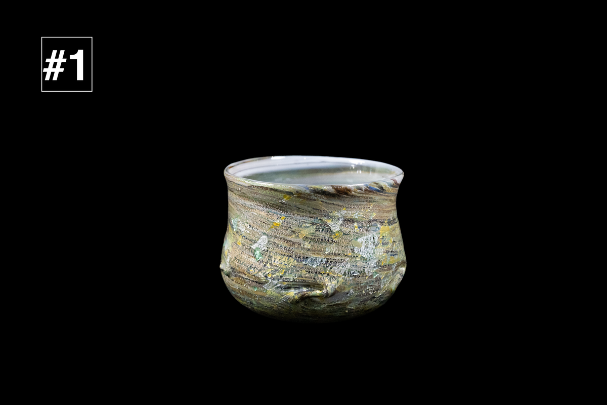 Handmade glass drinking cup with a softly flared rim and layered gray, gold, blue, and white swirls against a black background