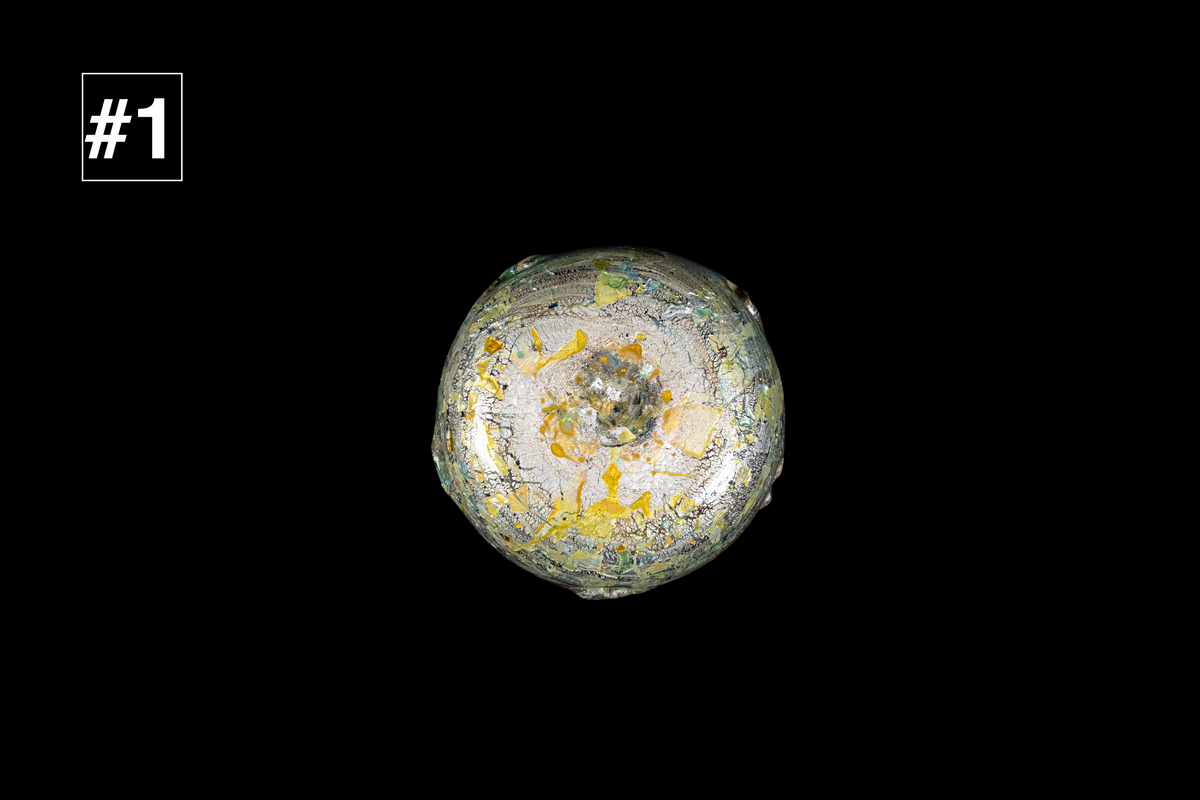 Bottom view of a handmade glass cup with swirling silver gray and gold flecks forming a rounded base against a black background
