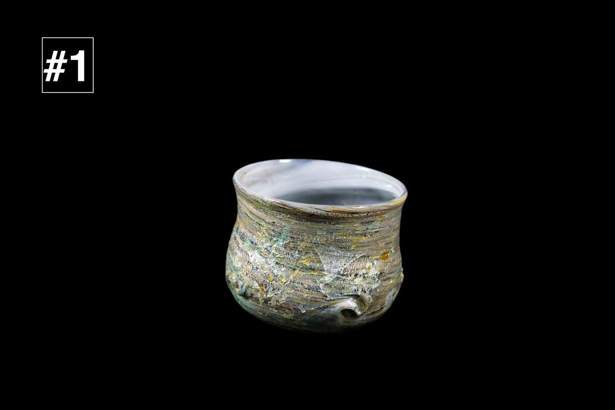 Small handmade glass drinking cup with a rounded shape, rippled multicolored texture, and pale blue curved rim against a black background