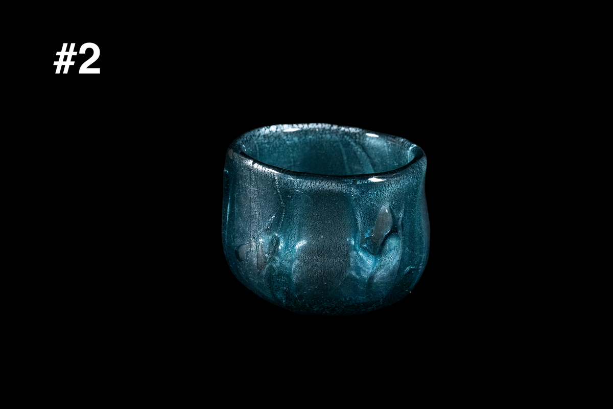 Small teal glass guinomi with a softly irregular rim and shimmering textured surface, handmade in a northern Japan studio against a black background