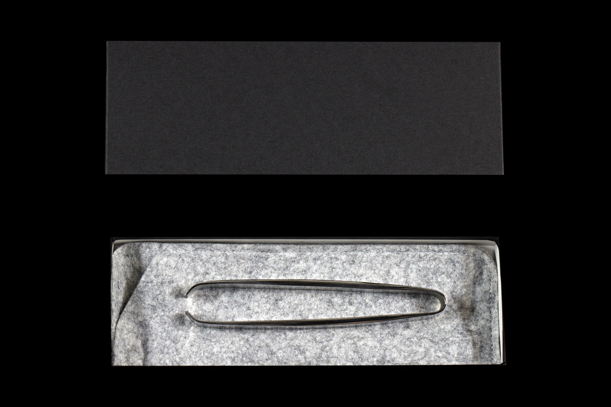 Hand forged stainless fish tweezers with a narrow tip resting on gray felt in an open box, with the dark gray lid above, on a black background