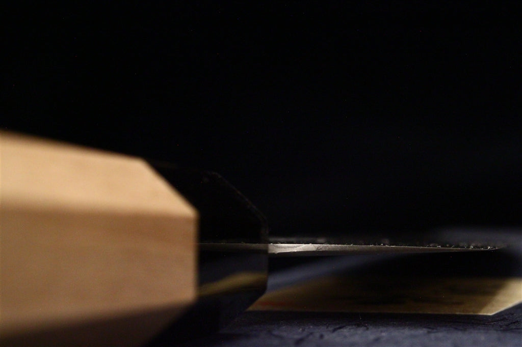 Side profile of a thin polished carbon steel gyuto blade with a light octagonal ho wood handle by Ashi Hamono against a black background