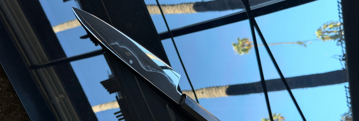 Mirror-polished Japanese kitchen knife with a dark octagonal handle, angled across a glass surface reflecting blue sky, palm trees and window frames