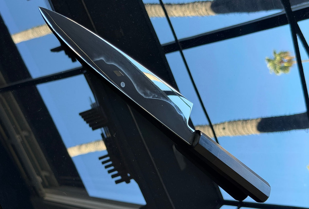 Close view of a mirror-polished Japanese kitchen knife with a dark octagonal handle, resting on glass that reflects blue sky and palm trees